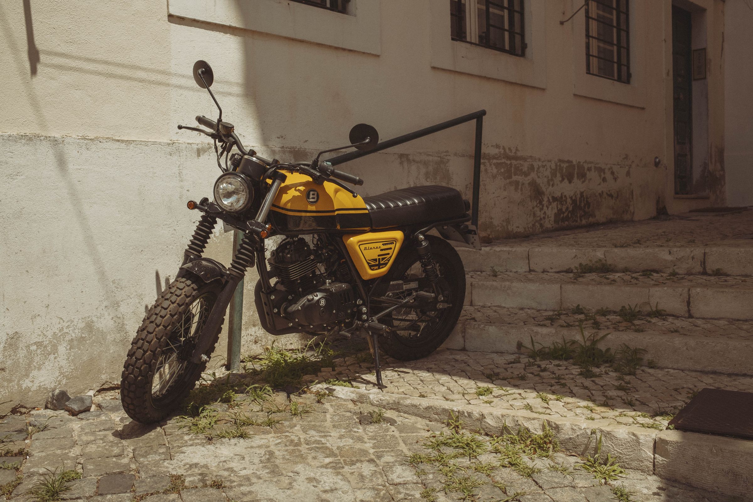 A yellow and black vintage motorcycle with knobby tires sits on its kickstand against a weathered two-tone wall, weeds growing through the cobblestones beneath it. The scrambler-style bike with its ribbed seat and round headlight is bathed in afternoon light, shadows falling across the quiet residential street.Claude is AI and can make mistakes. Please double-check responses.