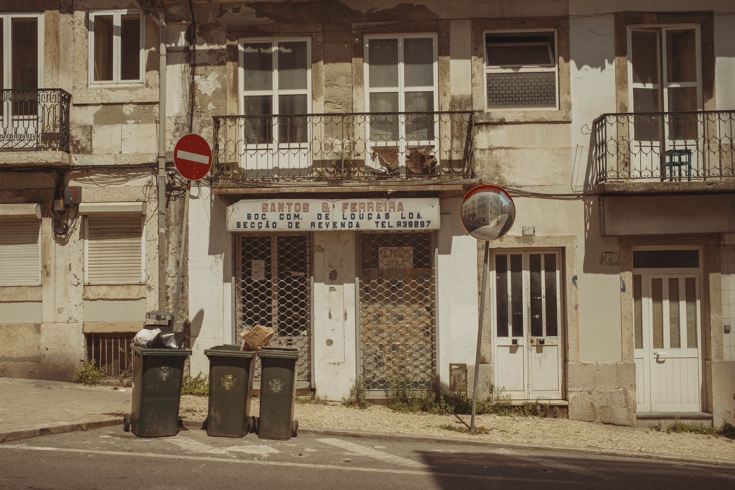 A derelict storefront with faded lettering reading 'SANTOS & FERREIRA SOC. COM. DE LOUÇAS LDA.' sits shuttered behind metal grates, its weathered facade peeling and stained. Three trash bins line the sidewalk in front of the abandoned building, while a no-entry sign and convex mirror mark the quiet street corner, iron balconies rusting above.
