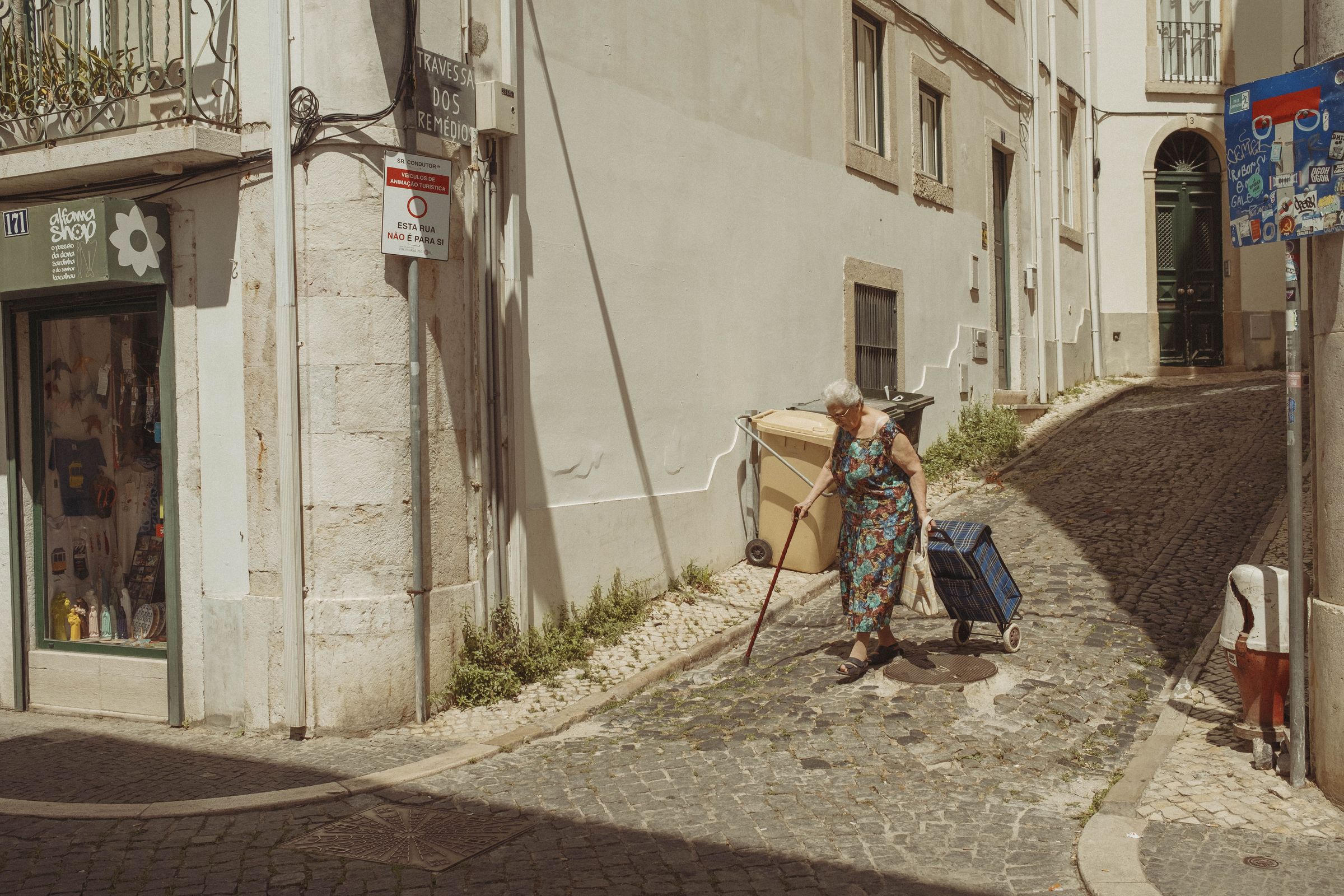 An elderly woman with white hair pulls a plaid shopping cart up a steep cobblestone street, using a cane for support as she climbs past whitewashed buildings. Weeds grow between the stones while shop signs and power lines line the quiet residential lane in the bright afternoon sun.