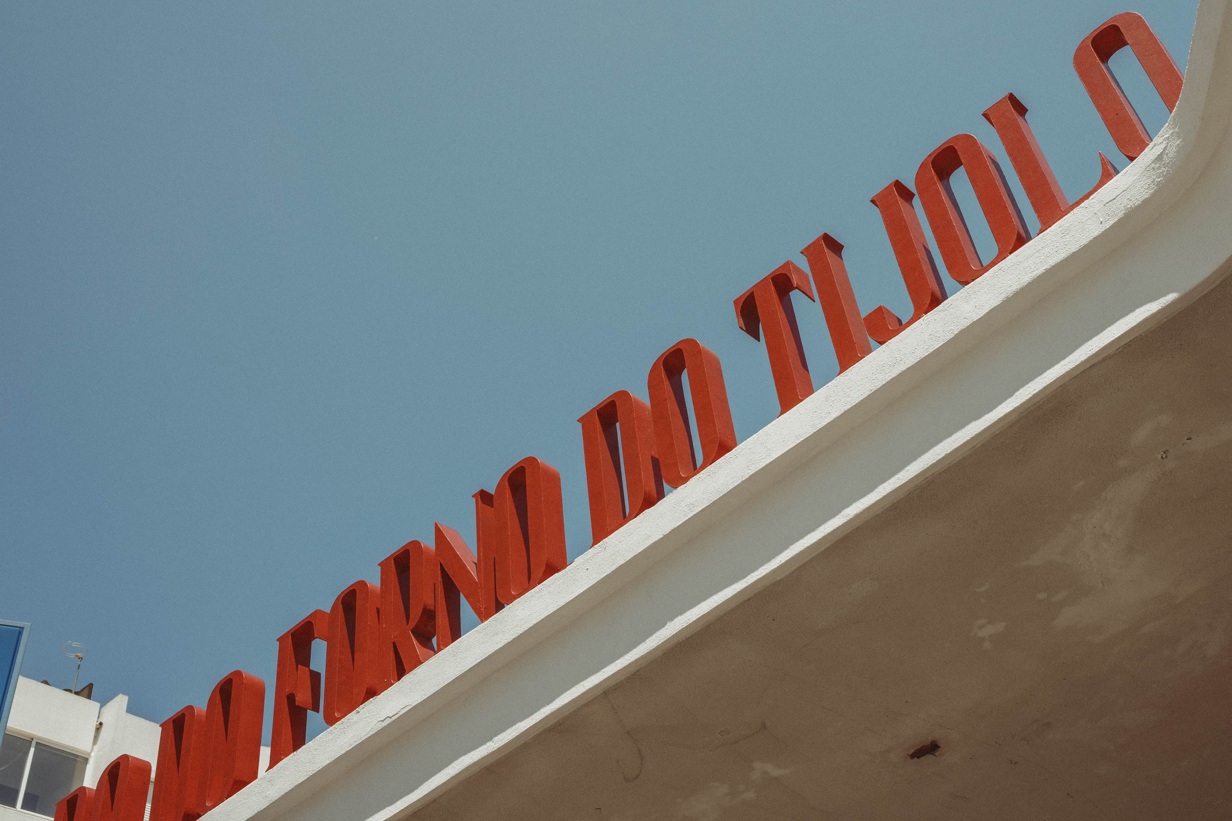 Large red dimensional letters angle upward along the edge of a modernist concrete roofline against a clear blue sky. The bold typography and stark color create a striking graphic composition with the building's clean white edge.