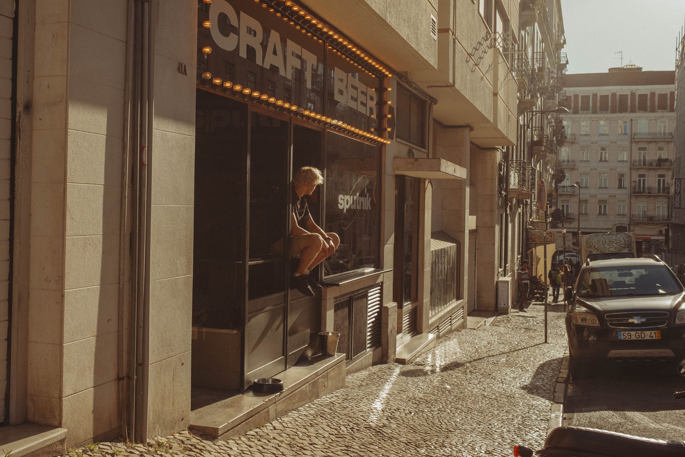 A person sits in the window of a craft beer bar with glowing marquee lights, looking at their phone as golden afternoon sun illuminates the cobblestone street. The urban scene shows pedestrians and a parked car on the sloped lane, with an ornate iron monument and apartment buildings rising in the background.