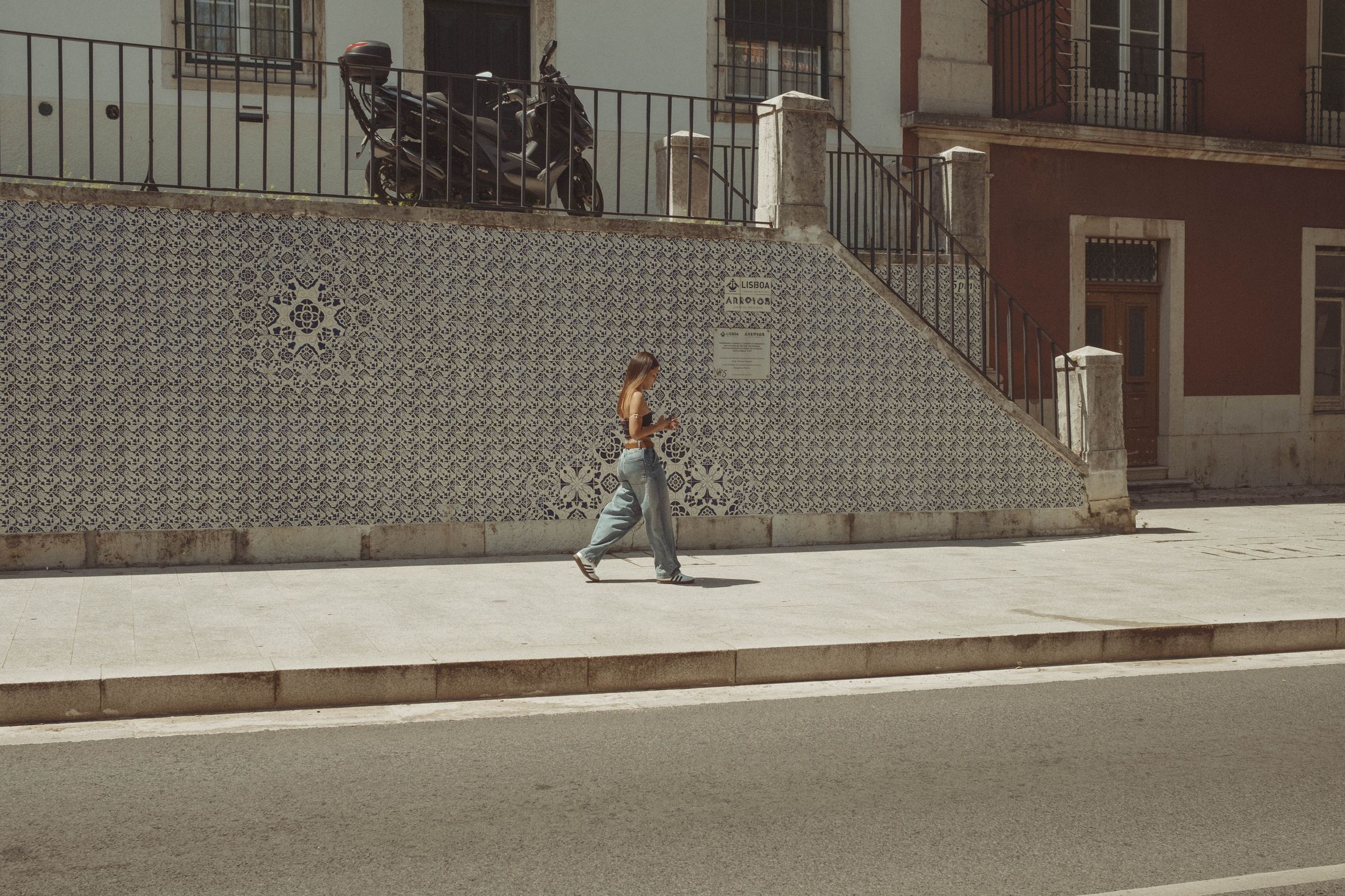 A woman in jeans walks across an empty sun-bleached plaza, checking her phone as she passes a wall covered in blue and white patterned azulejo tiles. A parked bicycle sits atop the stone steps behind her, with iron railings and weathered buildings framing the quiet urban scene.