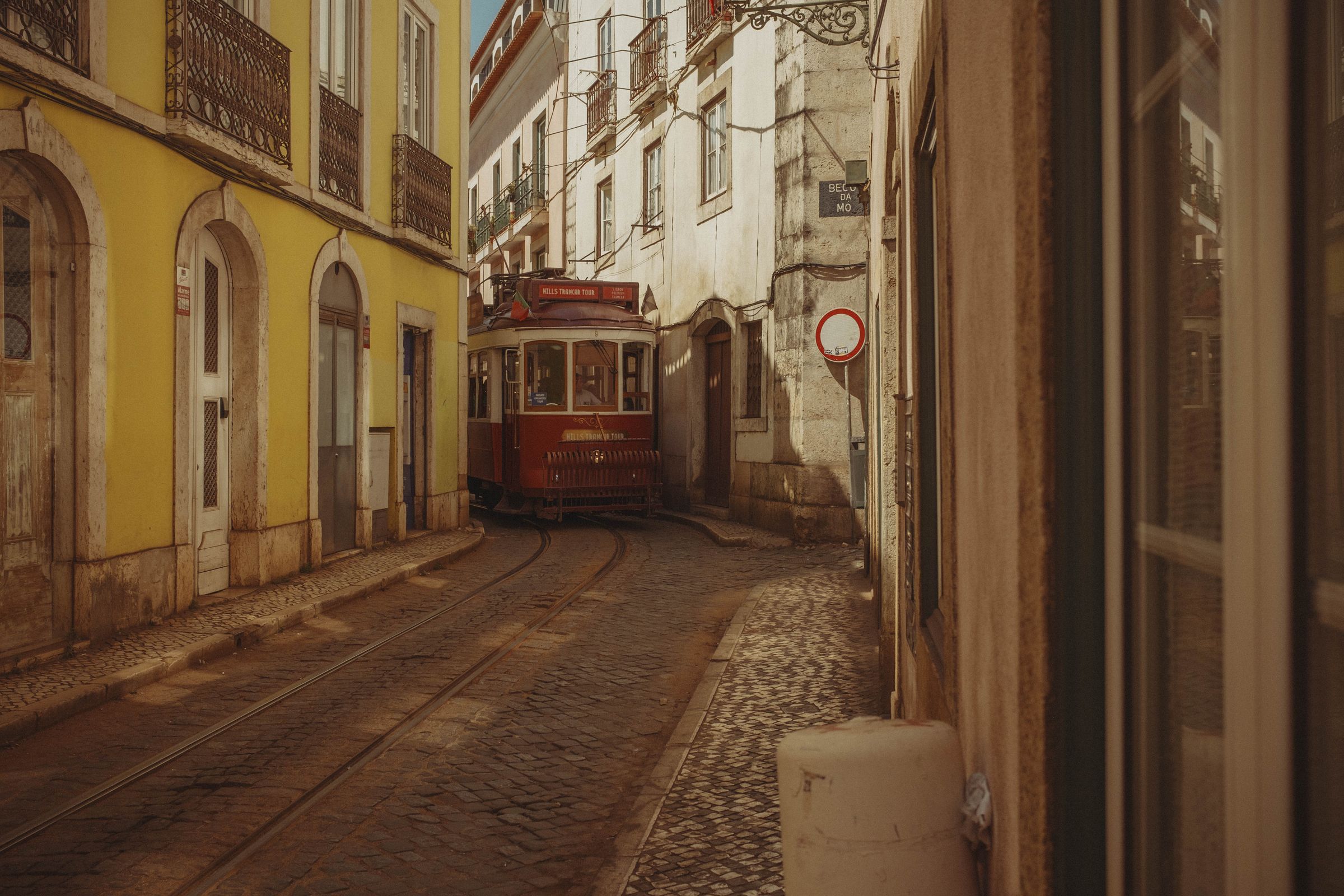 A vintage red tram navigates a tight curve on tracks through a narrow street lined with buildings in yellow, white, and weathered cream. The historic streetcar barely fits between the arched doorways and iron balconies, with overhead wires strung above and a no-entry sign visible on the stained wall ahead.