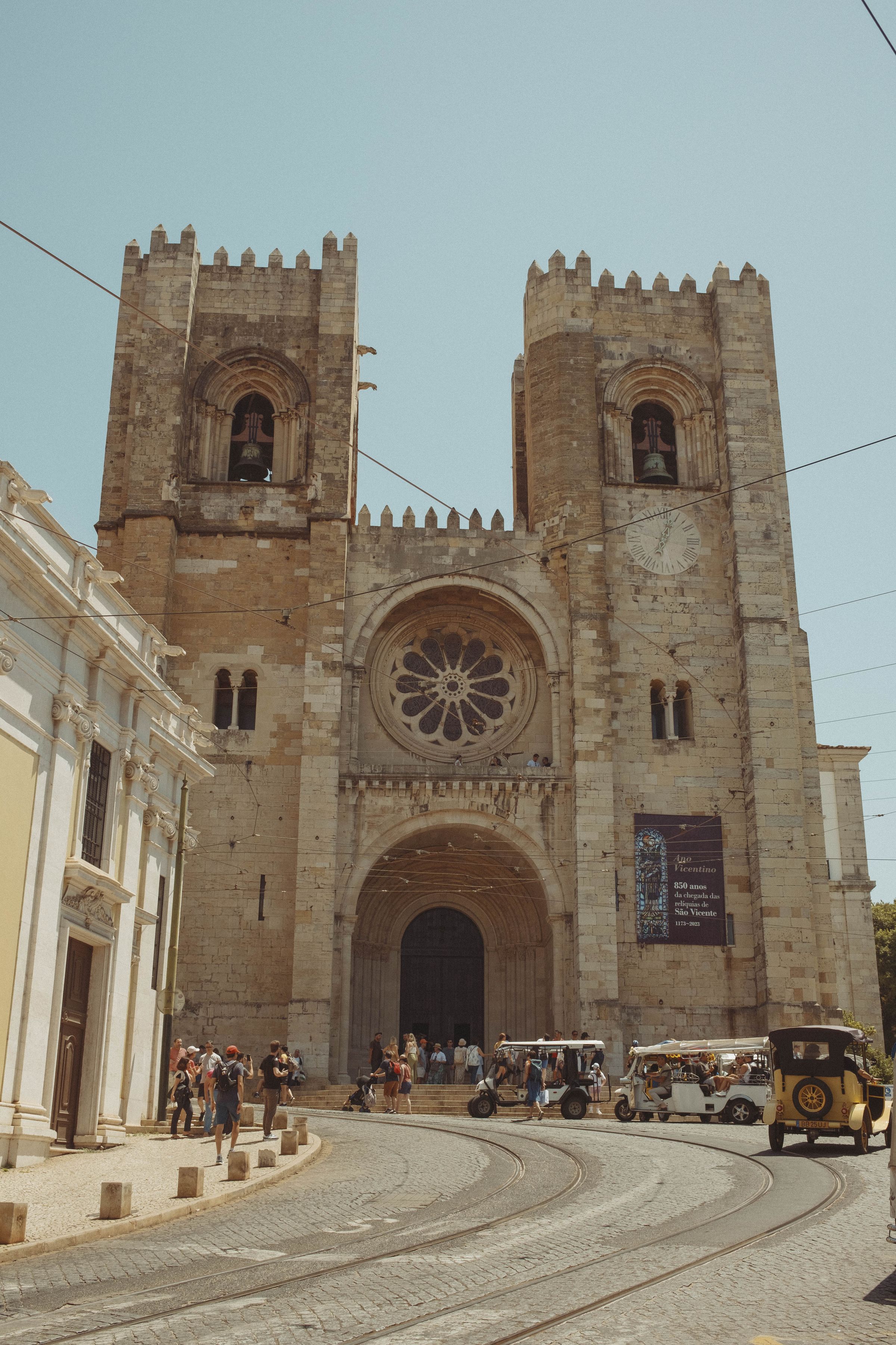The Sé de Lisboa cathedral rises with its twin crenellated towers and Romanesque facade, its rose window and arched portal dominating the plaza below. Tourists gather on the steps while tuk-tuks line up on the cobblestones, tram tracks curving past the ancient limestone structure under a pale blue sky.