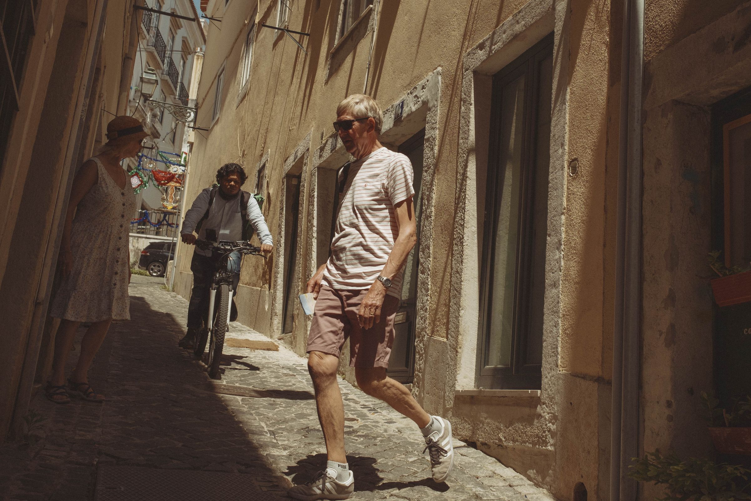 A man in sunglasses and striped shirt walks briskly up a narrow cobbled alley in harsh sunlight, while a cyclist and another pedestrian navigate the tight space behind him. The steep lane is lined with weathered tan and brown facades, their stone doorframes and worn plaster catching the afternoon light.