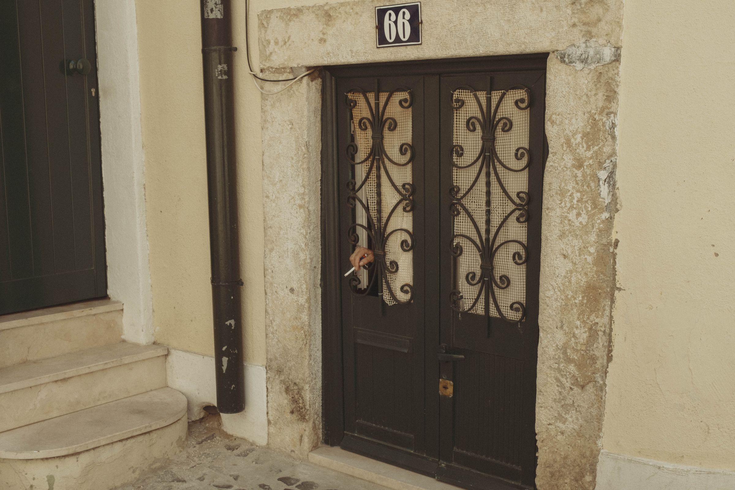 A dark wooden door with ornate wrought-iron scrollwork and mesh panels sits at number 66, a simple padlock securing the weathered entrance. The doorway is framed by aged stone and cream plaster, with worn steps leading to the neighboring building in the quiet residential passage. A hand holding a cigarette sticks out the window.
