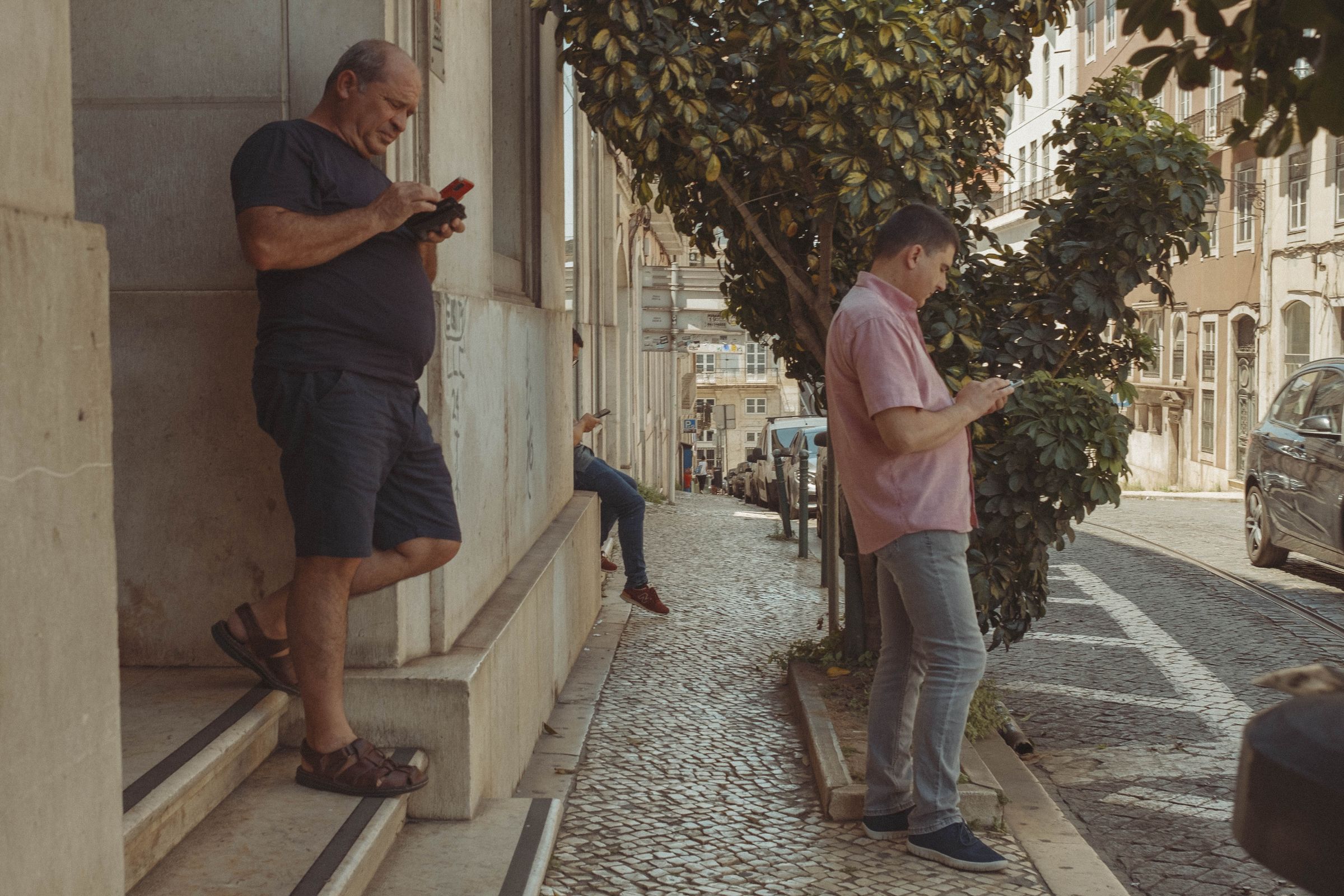 Three men stand absorbed in their phones on a sloping cobblestone street, one leaning casually against building steps while another stands beneath a tree. The scene looks down toward buildings cascading down the hillside, with parked cars and warm afternoon light filtering through the foliage.