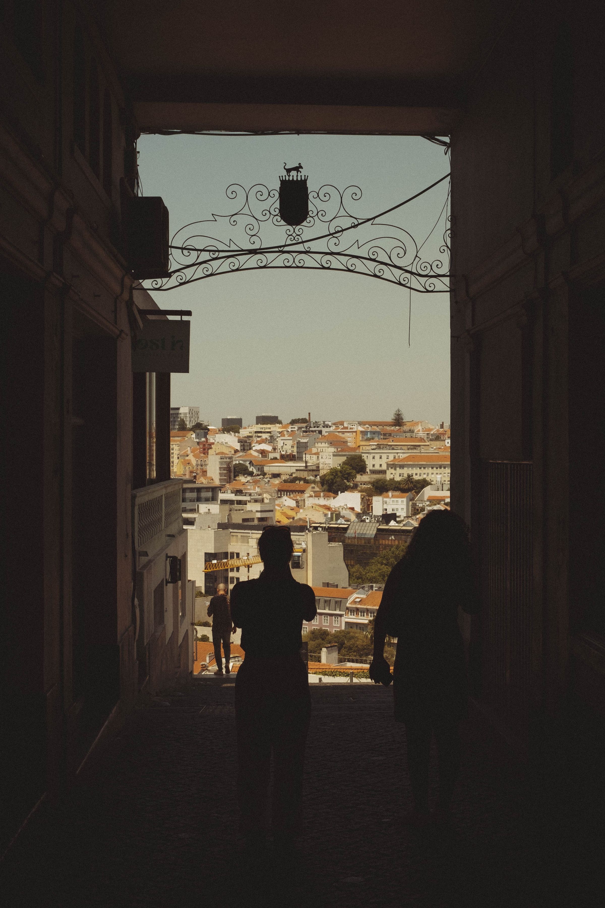 Silhouetted figures stand in a dark archway topped with ornate ironwork scrolls and a crown, framing a sunlit view of terraced buildings with terracotta roofs cascading down the hillside. The dramatic contrast between shadow and light creates a portal effect, drawing the eye from the enclosed passage to the sprawling cityscape beyond.