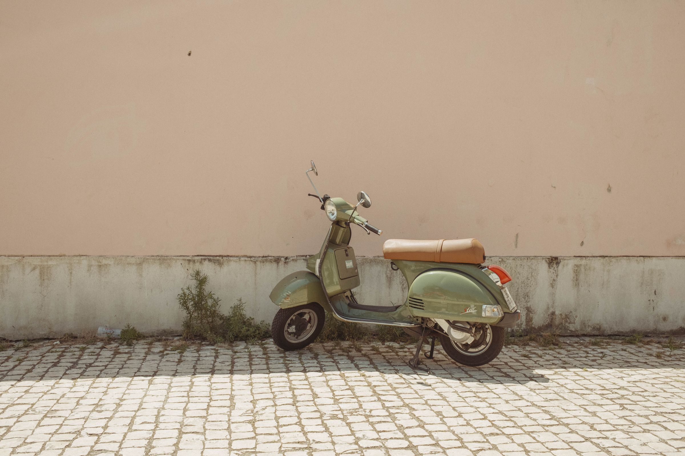 A mint green Vespa scooter with a tan seat sits on its kickstand against a pale pink stucco wall, weeds growing from the cobblestones at its base. The minimalist composition emphasizes the classic scooter's curves and chrome details against the weathered two-tone wall in soft afternoon light.