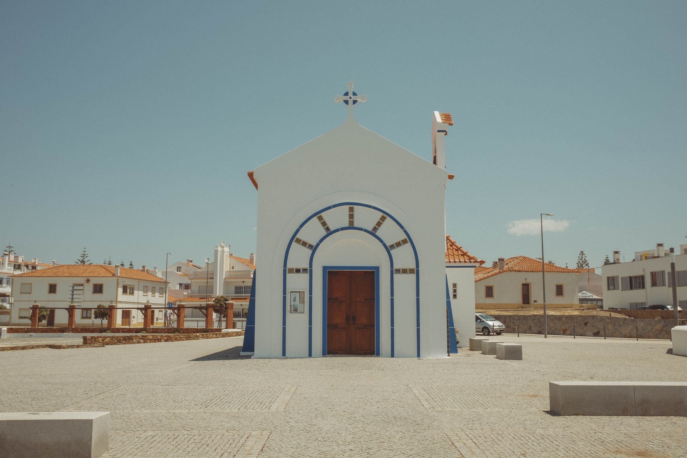 A small whitewashed chapel with blue trim stands alone in a sun-bleached plaza, its distinctive arched entrance decorated with decorative tile work and topped with a cross. The simple structure faces whitewashed houses with terracotta roofs under a clear blue sky, casting sharp shadows across the paved square.