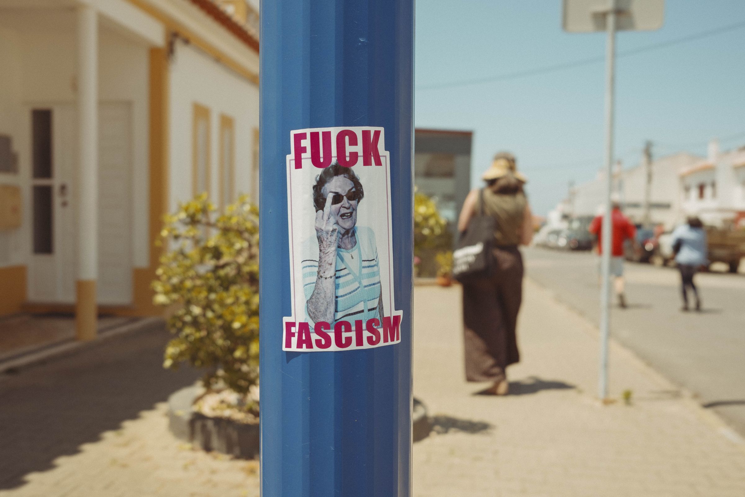 A sticker reading 'FUCK FASCISM' with an image of a person in sunglasses sticking up their middle finger is affixed to a blue pole on a sunny village street. Behind it, whitewashed buildings with yellow trim line the road where a blurred figure walks past in the bright afternoon light.