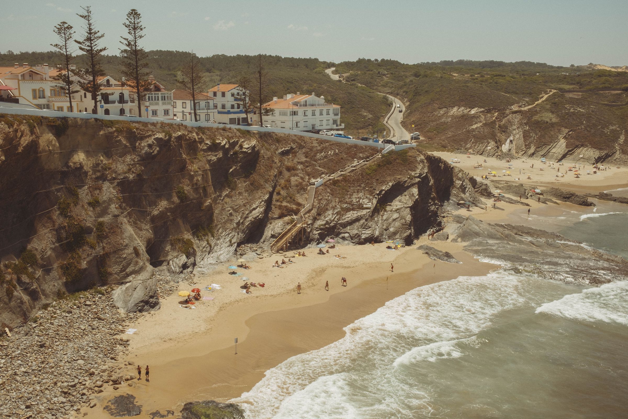 A dramatic cove beach nestles between towering dark cliffs, with beachgoers scattered across the sand and a wooden staircase descending the rock face. Whitewashed buildings with terracotta roofs perch atop the cliffs, while a winding road climbs through scrubland toward the horizon and waves roll onto the golden shore below.