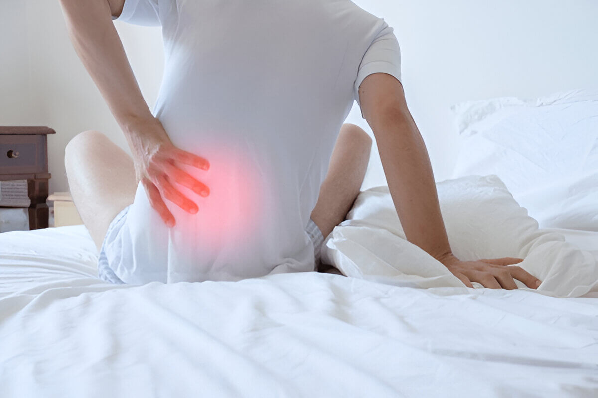 A man in pain with his arm on his lower back, sitting on the bed.