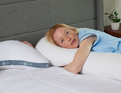 A woman lies on her side with arms wrapped around the Eli & Elm pregnancy pillow beside the side sleeper pillow