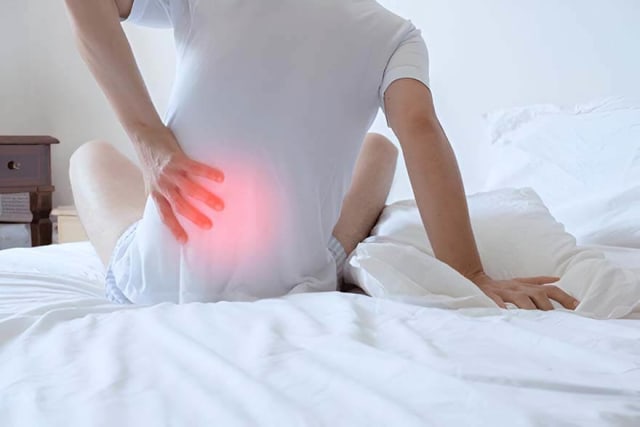 A man in pain with his arm on his lower back, sitting on the bed.