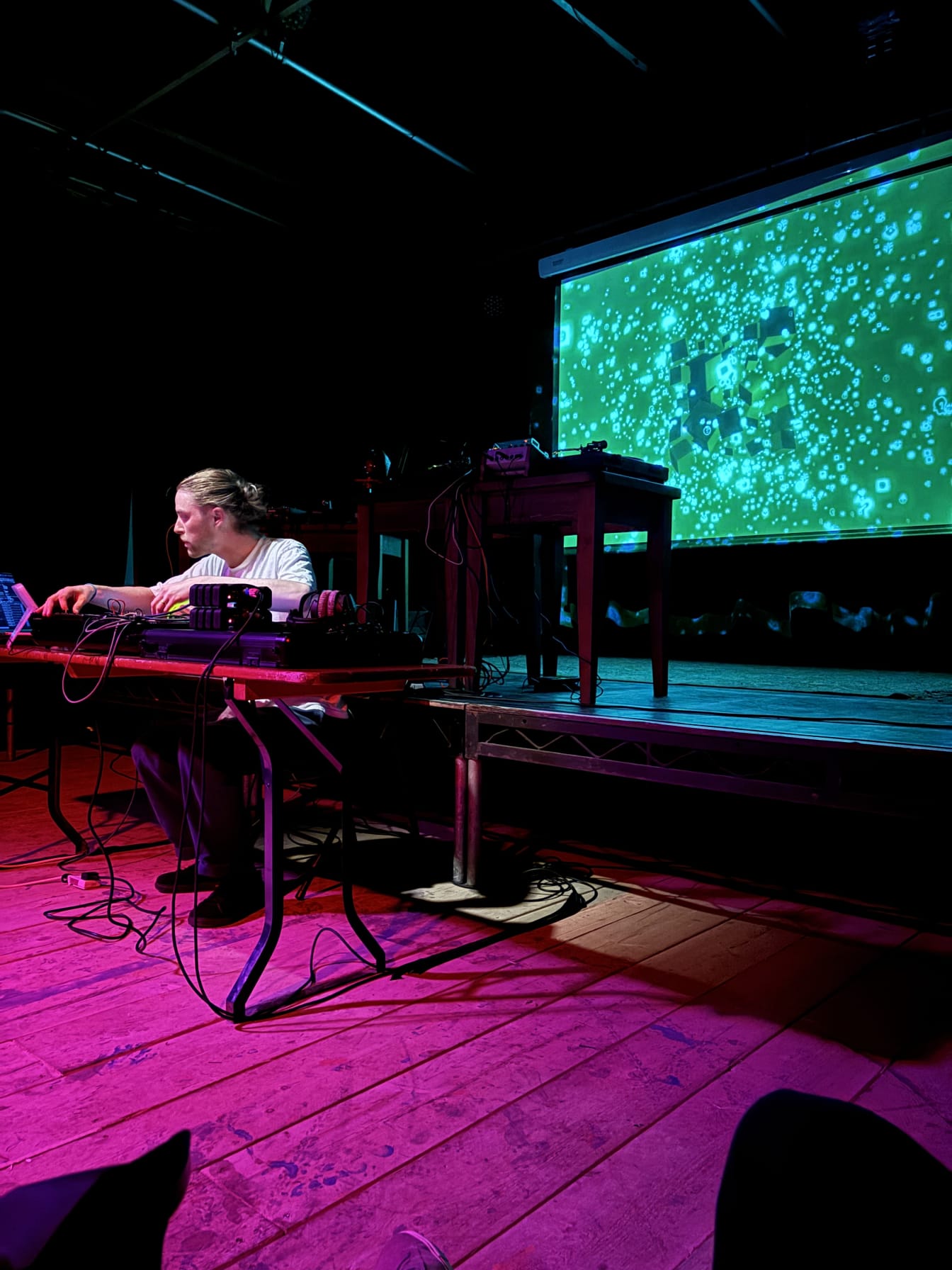 Musician Jonathan Fox playing experimental ambient music with various electronic equipment sitting at a desk in front of an audience, a projector cast behind the artist shows a cloud of emoji symbols on a green background