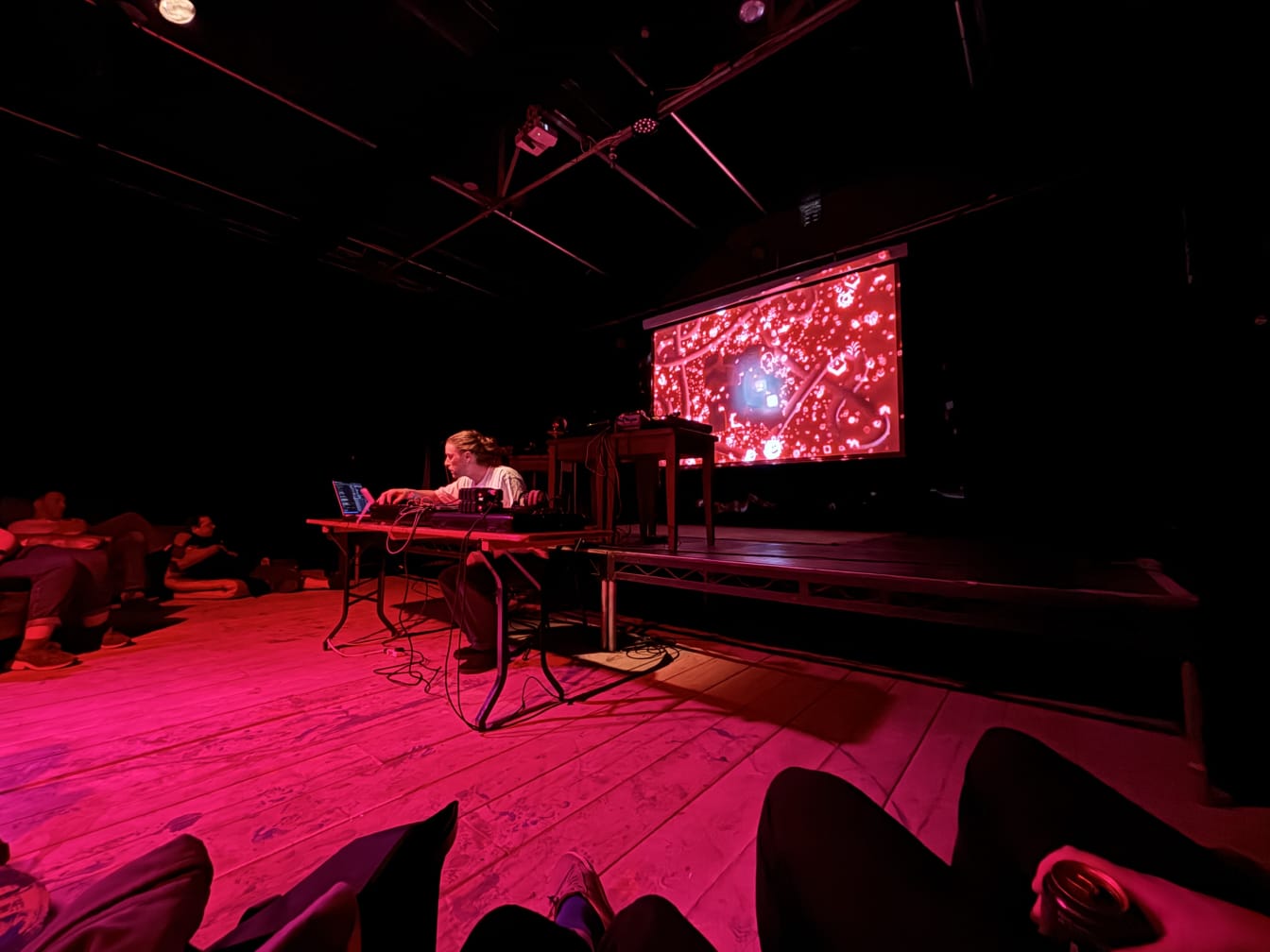 Musician Jonathan Fox playing experimental ambient music with various electronic equipment sitting at a desk in front of an audience, a projector cast behind the artist shows a cloud of emoji symbols on a pink background