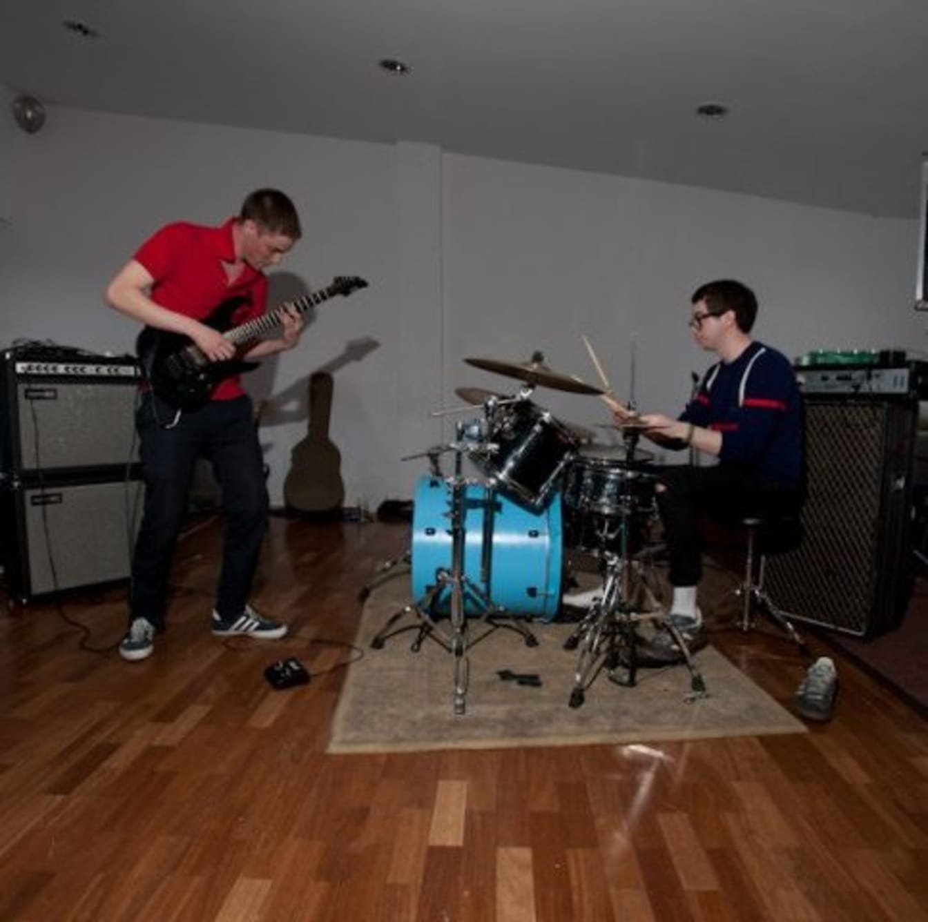 Two musicians play instruments in a practice room, with a guitarist standing and a drummer seated at a blue kit.