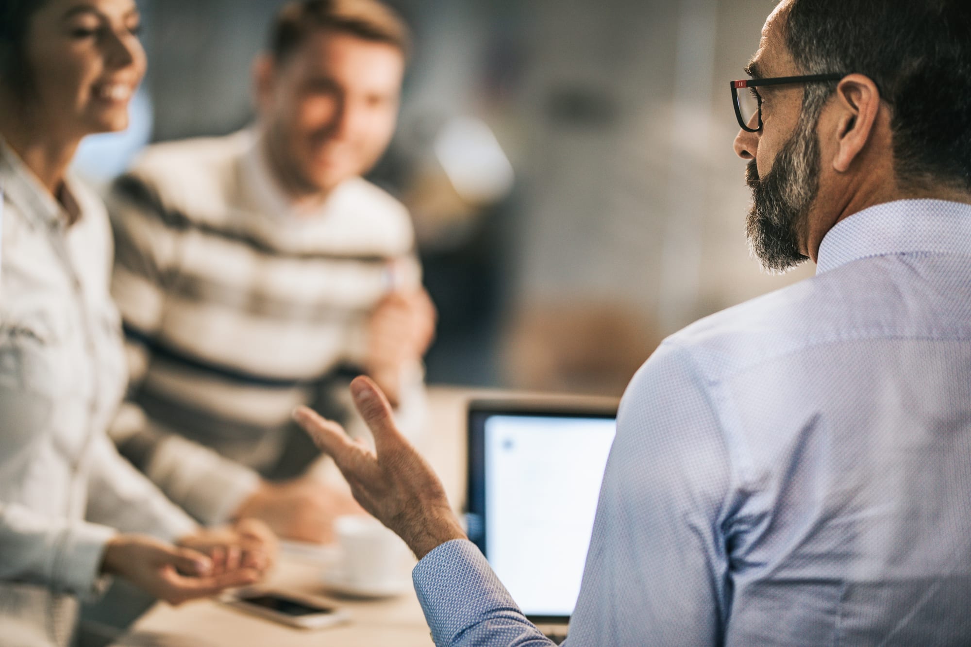 middle aged man speaking to a couple people in the background while sitting in front of a laptop and gesturing with his left hand