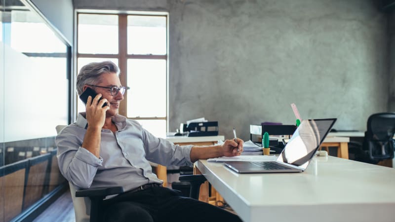 middle aged man in business attire smiling and writing on a piece of paper as he talks on a cell phone in front of a laptop