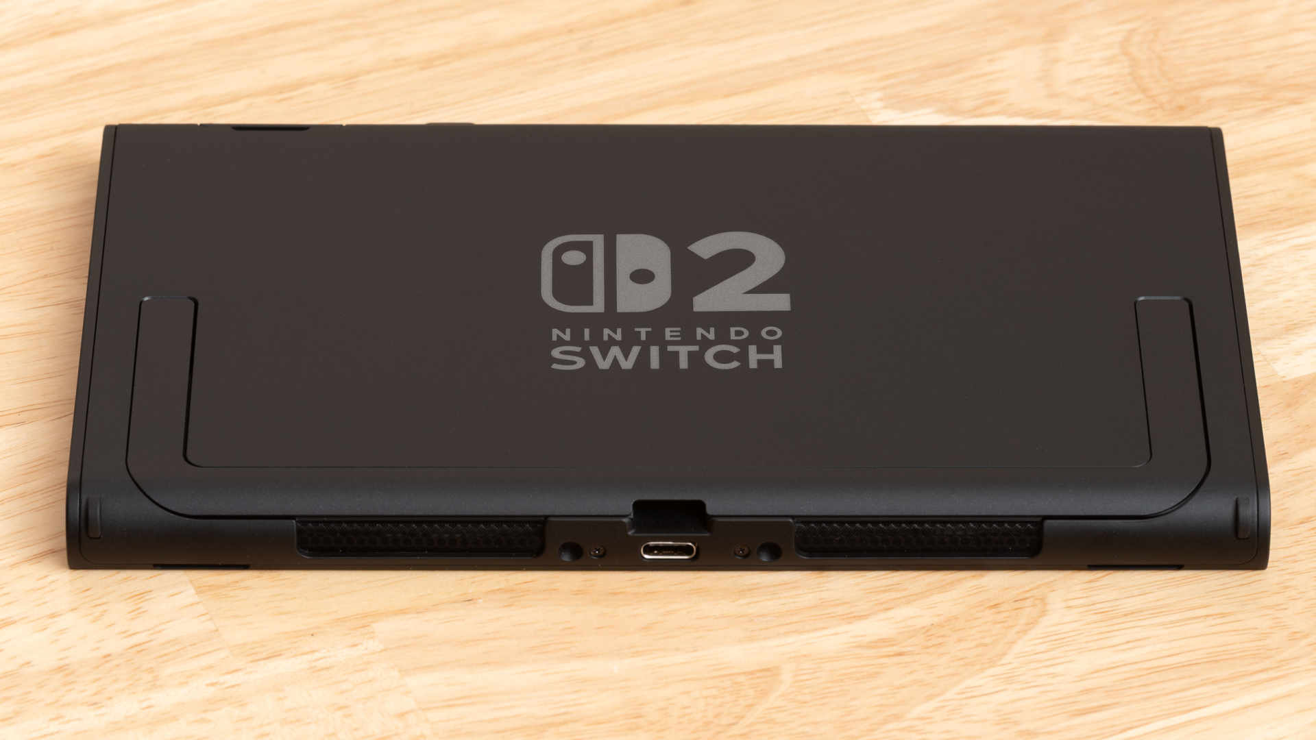 The back of a black Nintendo Switch 2 console lying flat on a wooden table, showing the Switch 2 logo, the folded kickstand and the bottom USB-C port