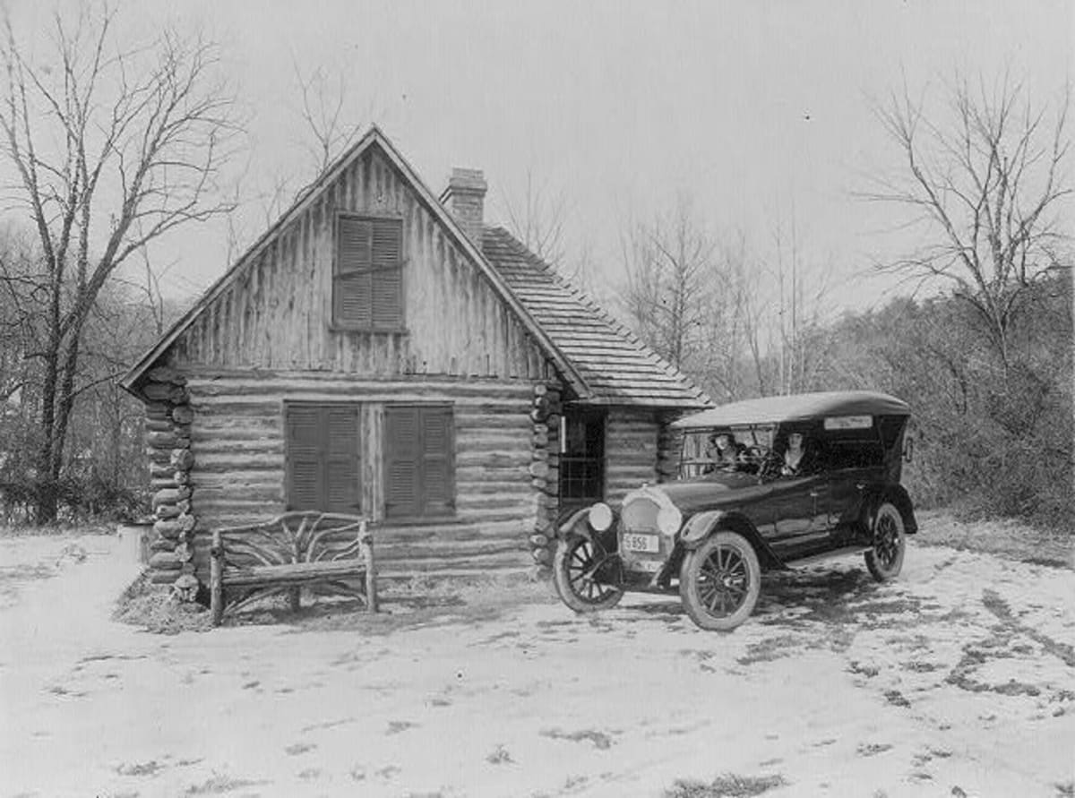Two women in a new open-top Oldsmobile touring car parked beside a rustic log cabin in Rock Creek Park, Washington, around 1920