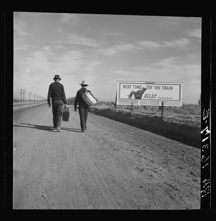 Two men walk along a California highway toward Los Angeles in 1937, passing a large billboard that reads Next time, try the train. Relax.