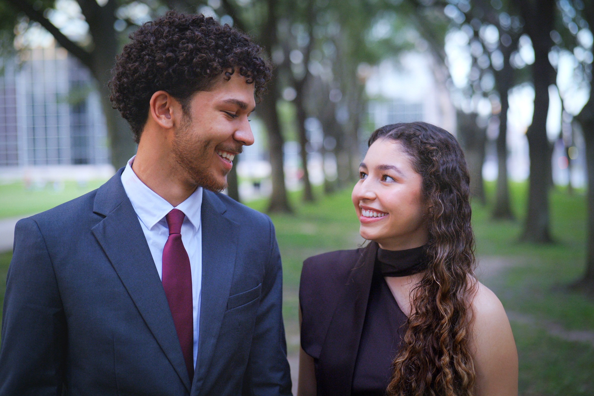 Couple in formal attire smiling at each other under the trees
