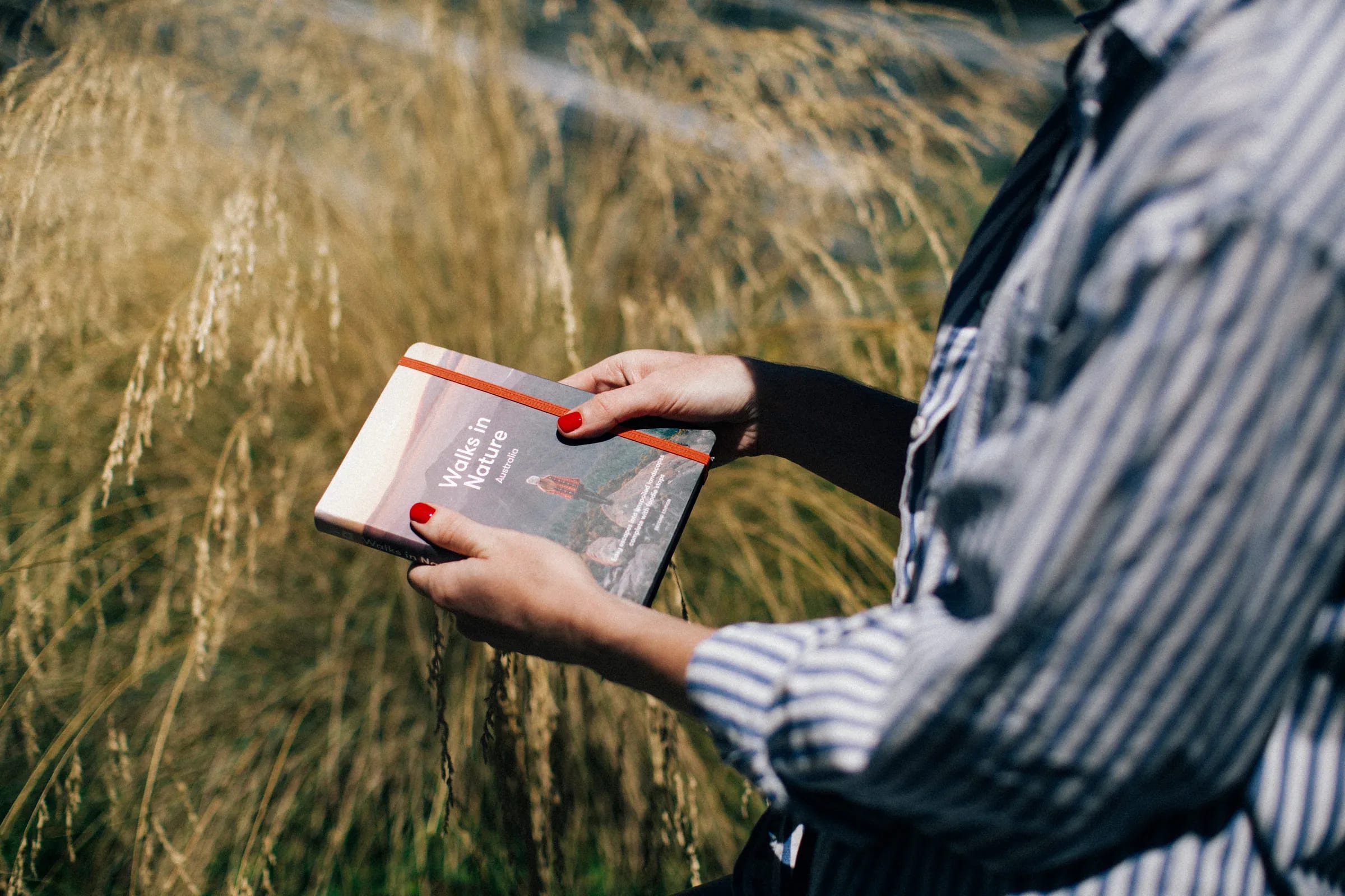 Hands with a red manicure hold small book with grassy background.