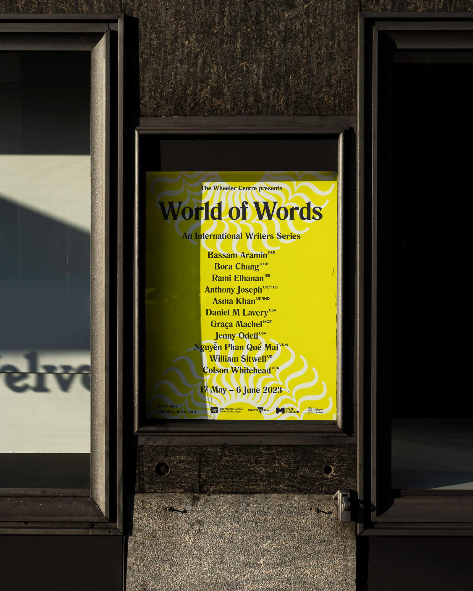 Yellow poster displayed on a wall slightly cast in shadow on the left hand side. Large text on the poster reads World of Words in back. Small black text sits below.