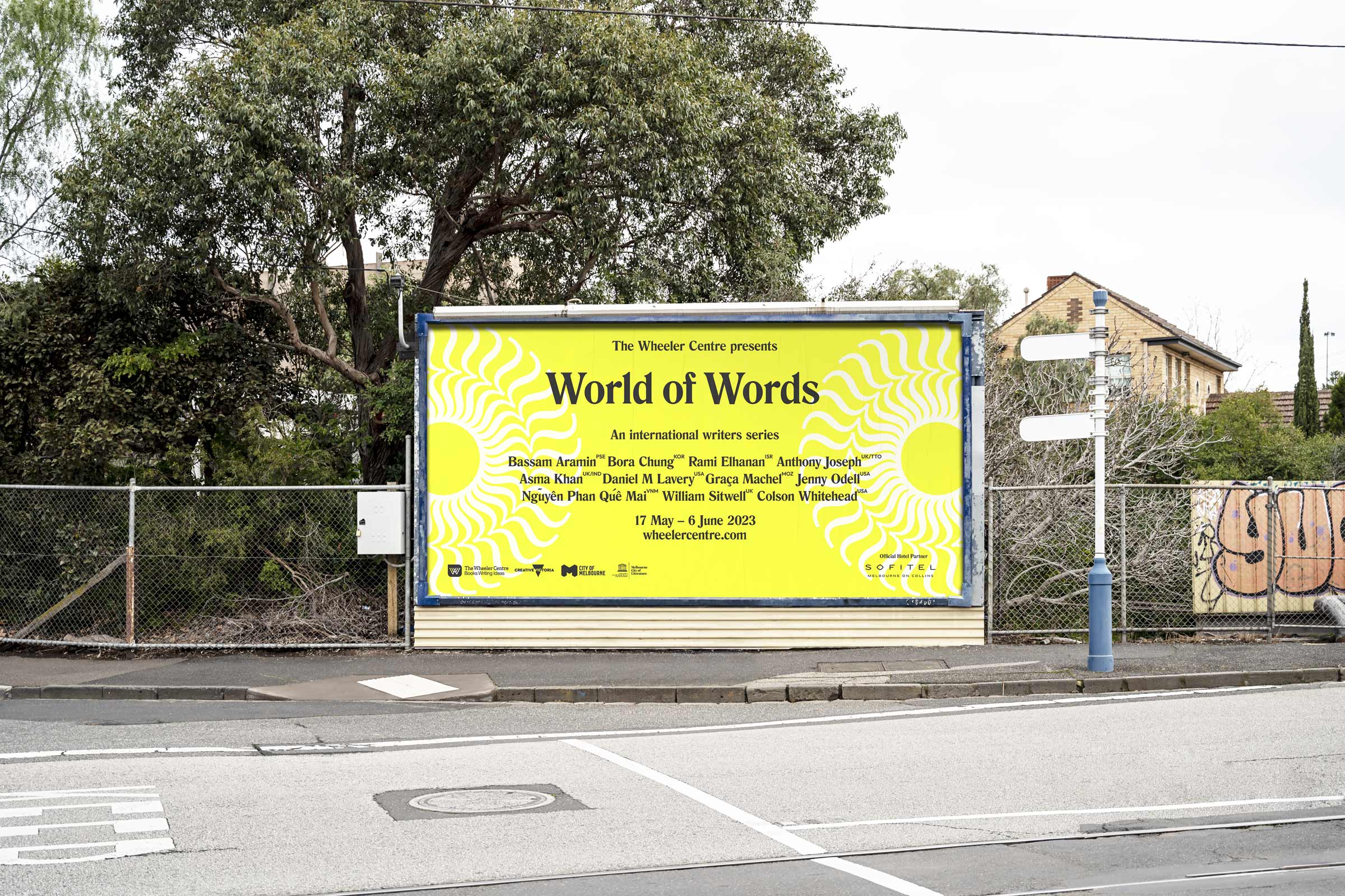 Large bight yellow billboard displayed on street surrounded by trees and fences. Black text on the billboard reads World of Words with smaller text below.