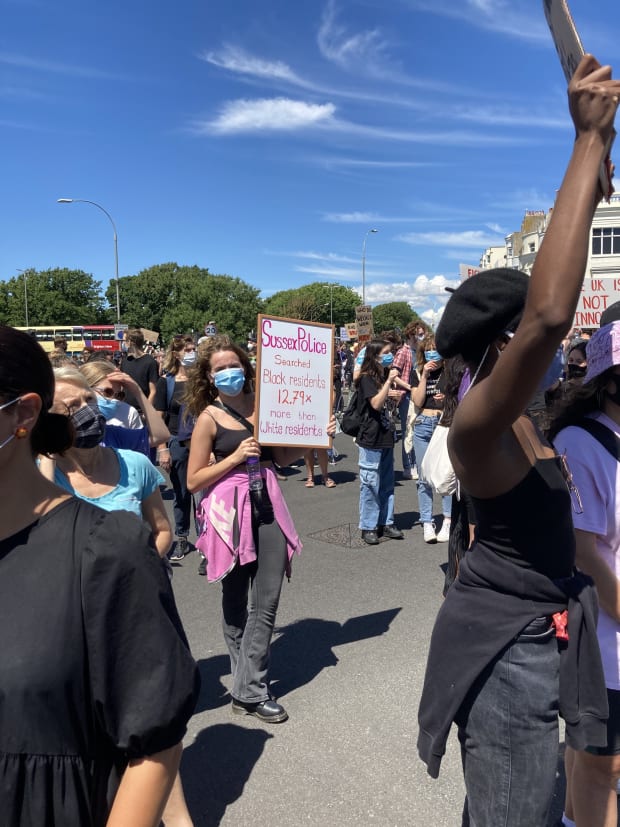 A woman standing in a crowd holding a sign saying Sussex police searched black residents 12.79x more than white residents