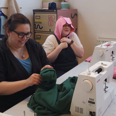 Two smiling Make Mee students sitting at their sewing machines. One of them has draped a partially-made hood over her head.