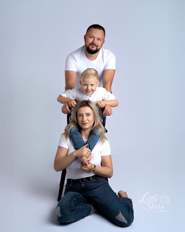 Rodzina trzech osób w białych koszulkach pozuje w układzie piramidy na jasnym tle.