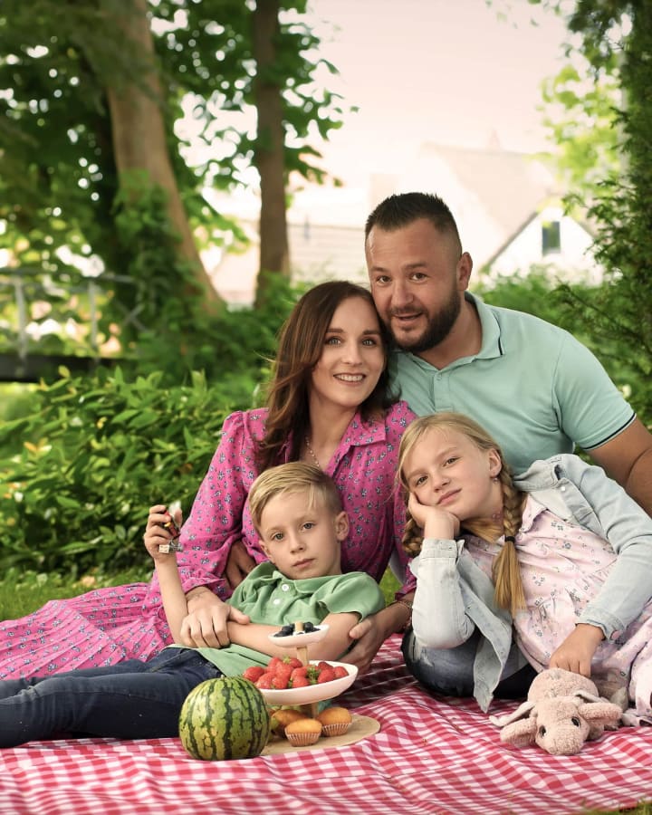 Rodzina siedzi na różowo-białej kraciastej tkaninie w ogrodzie, otoczona zielenią i kwiatami.