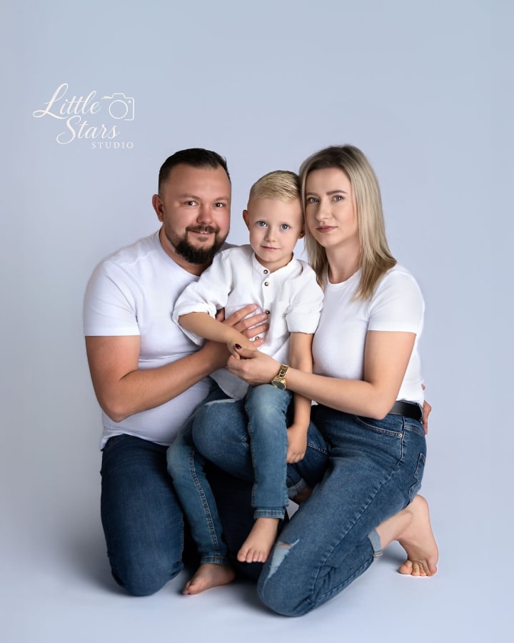 Rodzina trzech osób w białych koszulkach i jeansach pozuje na szarym tle, przytuleni do siebie.