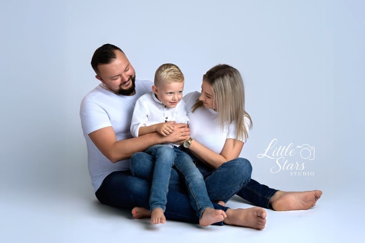 Rodzina siedzi na białym tle, mężczyzna i kobieta przytulają małe dziecko w białych koszulkach.
