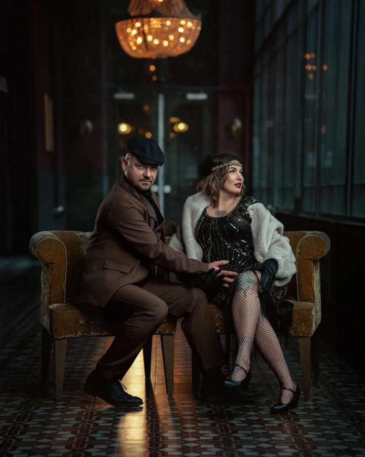 Para siedzi na złotej sofie w eleganckim wnętrzu, oświetlona ciepłym światłem lampy.