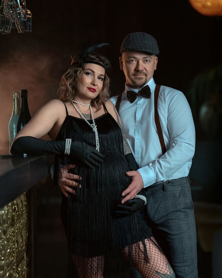 Para w stylu vintage czeka na dziecko, kobieta w czarnej sukience i długich rękawiczkach, mężczyzna w kamizelce i czapce.