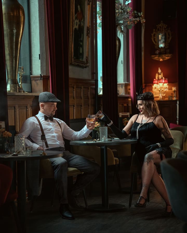 Para siedzi przy stoliku w eleganckim wnętrzu, mężczyzna w kamizelce i kapeluszu, kobieta w czarnej sukni.