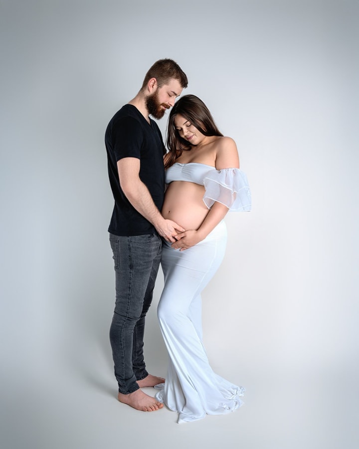 Para pozuje razem, mężczyzna obejmuje brzuch kobiety w ciąży, białe tło.