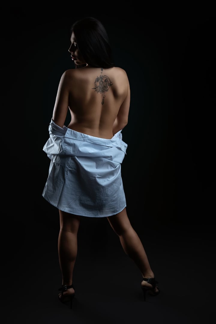 Woman with back tattoo in sensual portrait session, minimalist styling with shirt, dark background.