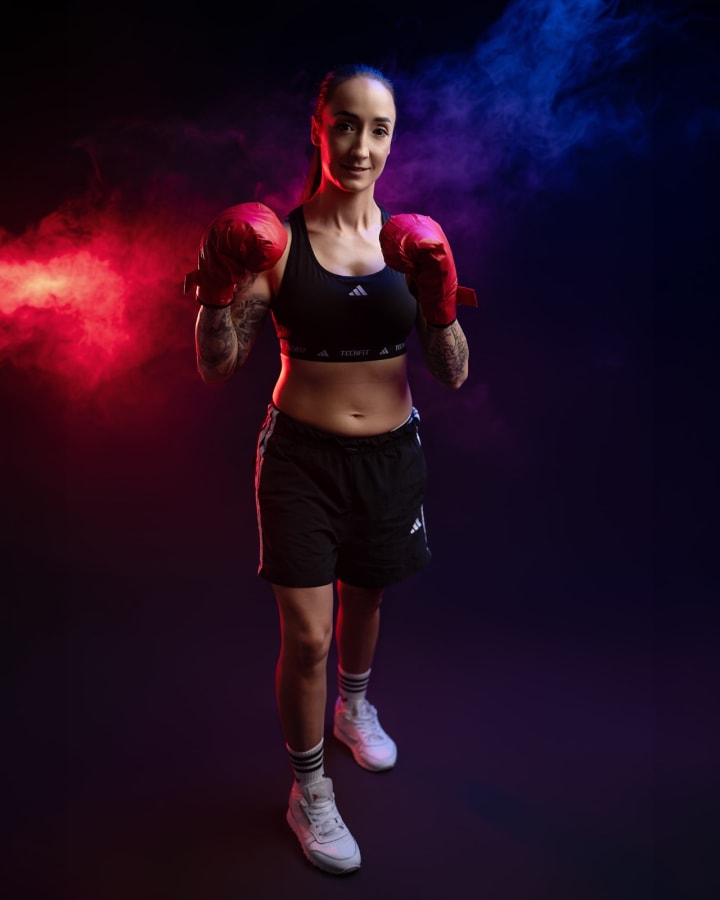 Kobieta w sportowym stroju i czerwonych rękawicach bokserskich na tle dramatycznego oświetlenia w odcieniach czerwieni i błękitu.