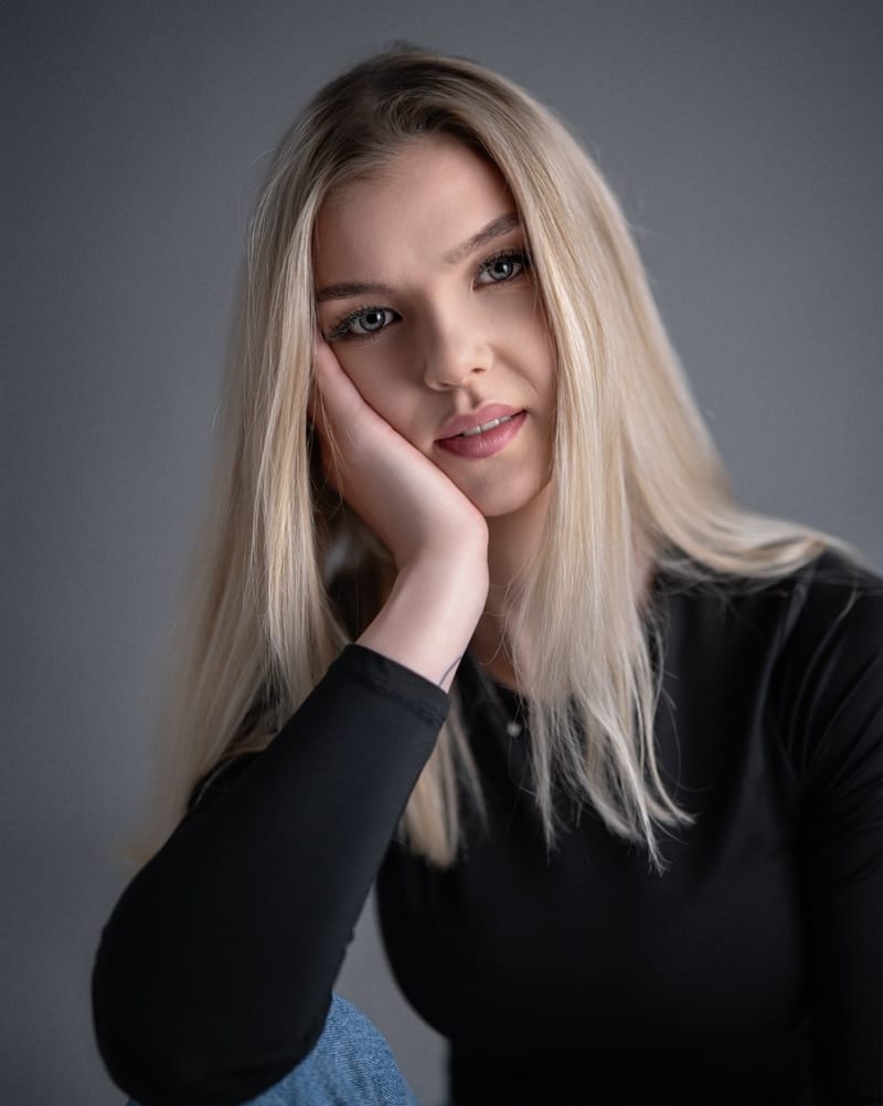 Portret nastolatki z długimi blond włosami w czarnej bluzce, dłonią przy twarzy, na jednolitym szarym tle.