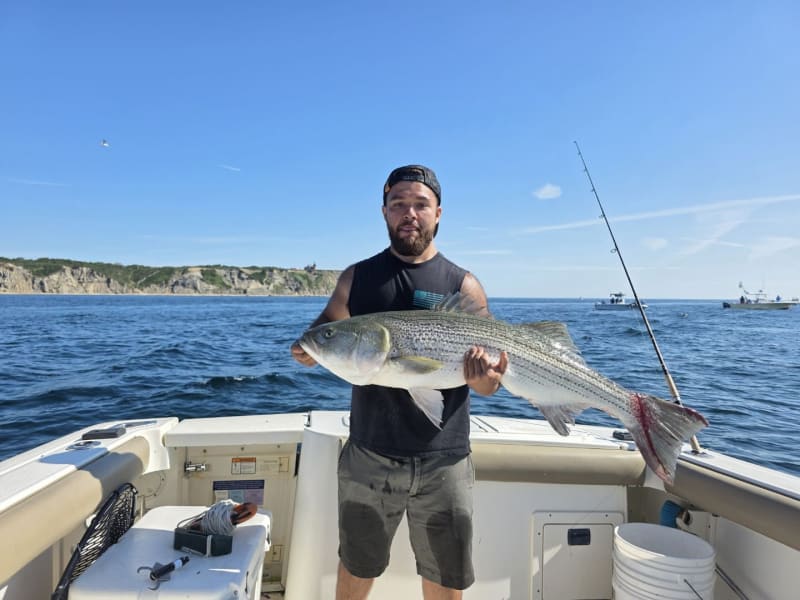 Angler in a black tank holding a large striped bass with the Block Island bluffs behind the Hula Dog