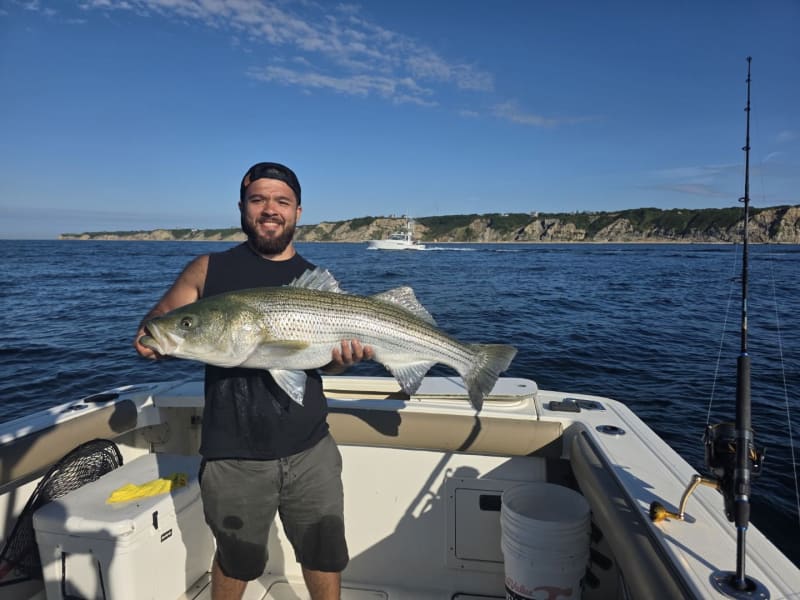 Angler holding a trophy striped bass off Block Island with the bluffs and a passing sportfishing boat behind