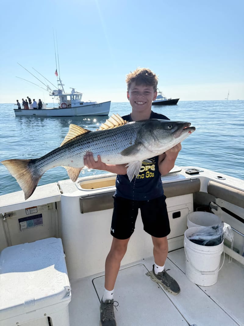 Young angler holding a large striped bass aboard the Hula Dog with harbor boats in the background