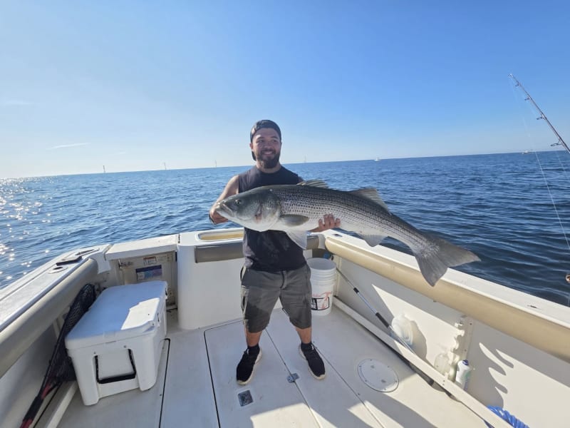Angler holding a striped bass aboard the Hula Dog with the Block Island Wind Farm on the horizon