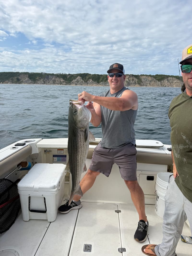 Angler holding a trophy striped bass aboard the Hula Dog with the Block Island bluffs behind