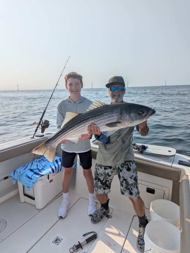 Kid and Captain Matt holding a big striper with the Block Island Wind Farm turbines visible behind them